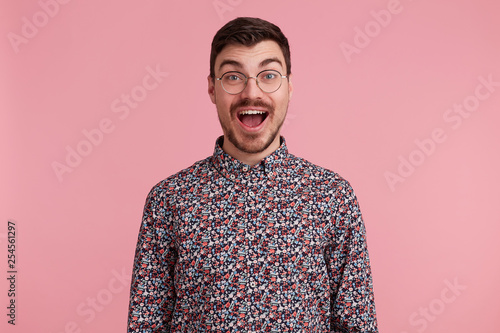 Portrait of a happy excited attractive handsome young man wearing glasses with dark hair unshaved with beard and mustache in colorful shirt opened mouth in amazement, isolated over pink background