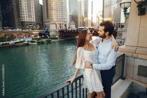 beautiful long-haired girl in summer dress with her handsome husband in white shirt and pants standing in sunny Chicago near river