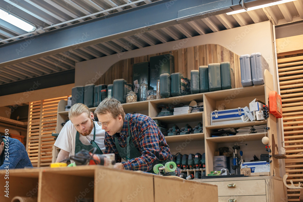 Φωτογραφία Two woodworker craftsmen are examining the drawings of the product of a new project while standing in their carpentry against the background of shelves with tools