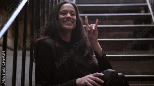 Medium shot of a beautiful mixed heritage woman sitting on some stairs outdoors and doing a rock on hand gesture while looking at camera