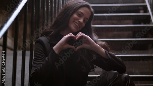 Medium shot of an attractive woman making a heart symbol both hands while sitting outdoors on some stairs