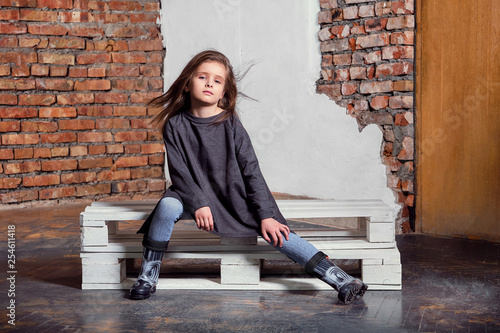 Little kid girl model posing fashionable in casual stylish clothes,  gumboots. Fashion child sitting pose. studio background, brick wall.Shop  youth, advertisement. Stock Photo