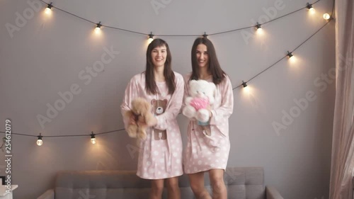 Two beautiful female twins with teddy bear dancing joyfully and smiling on gray background in slowmotion photo