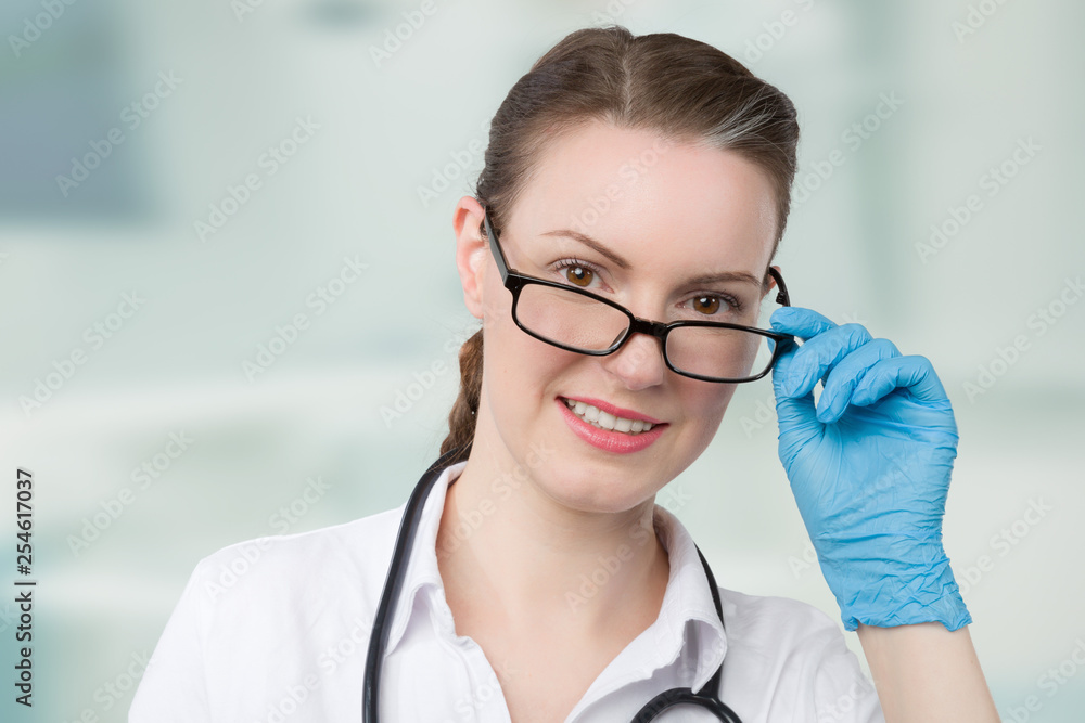 young doctor with blue medical gloves looks over her glasses in front of a clinic room young doctor with blue medical gloves looks over her glasses in front of a clinic room