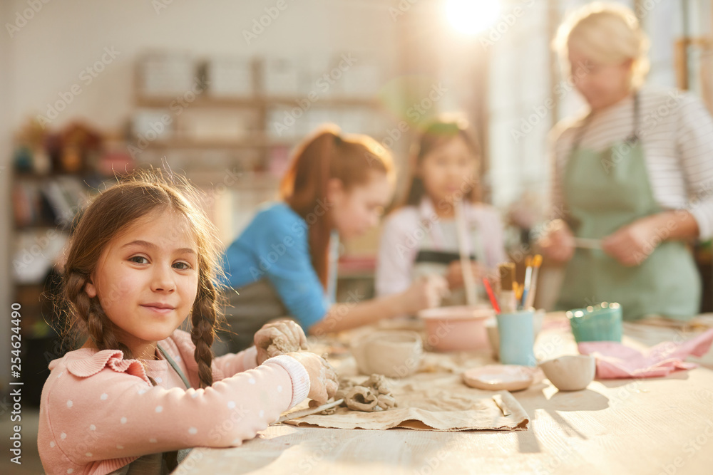 Portrait of cute little girl looking at camera while enjoying pottery class with group of children, copy space