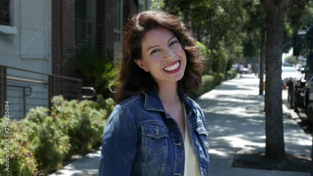 Woman smiles and laughs into camera on sidewalk.