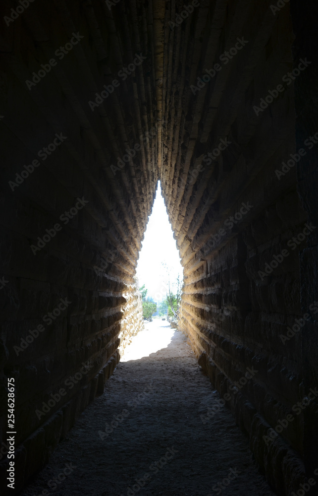 Path through a dark narrow tunnel in old stone ruins. Sunlight and green trees at the end of the tunnel