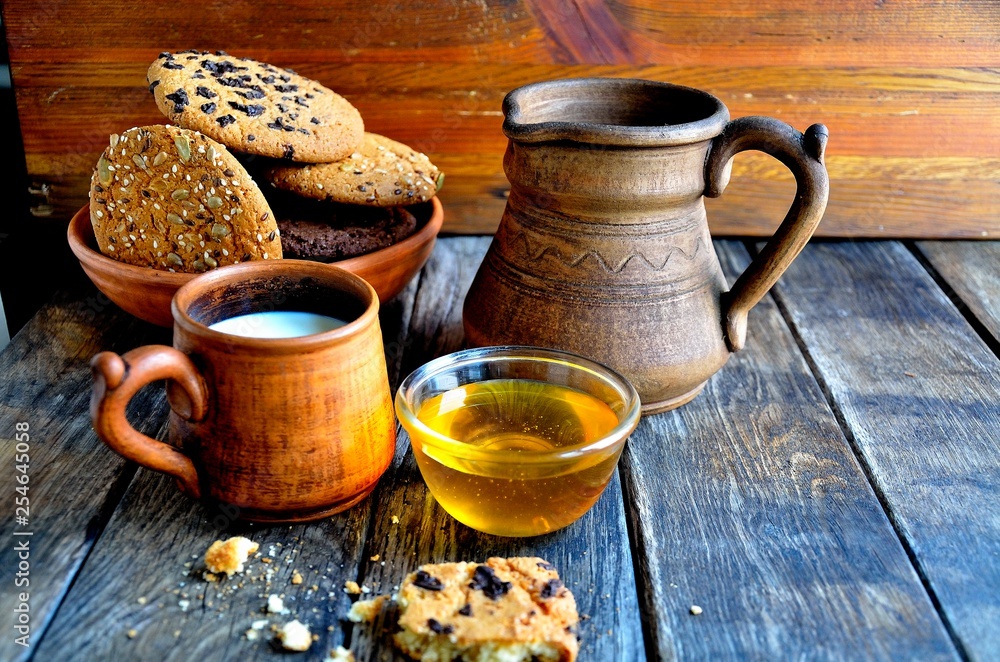 Oatmeal cookies in a ceramic rustic plate, milk in a ceramic mug, napkin, honey on a rustic wooden table.