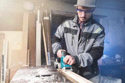 Young carpenter with a beard working with an electric plane with suction of sawdust. Leveling and sanding wooden bars