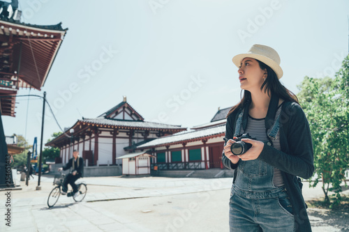 Beautiful young asian lady tourist at traditional japanese temple shitennoji in osaka japan. girl standing under blue sky hold camera photographing while traveling. male in suit riding bike bicycle.