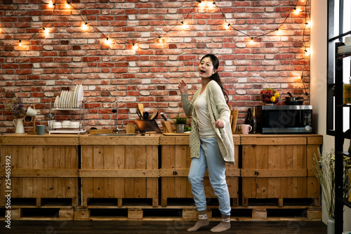 Cheerful young funny asian woman dancing and singing while having leisure time in kitchen at home. warm winter modern place apartment with red brick wall and hanging up light. elegant female relax.