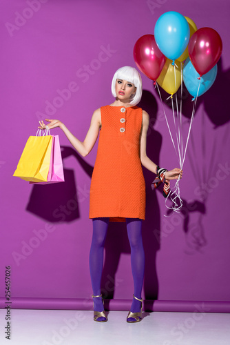 Sad girl in white wig holding shopping bags and air balloons on purple background