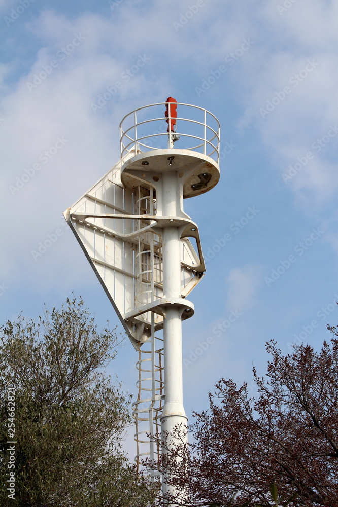 Lighthouse beacon warning light on top of strong metal structure surrounded with safety fence and tall trees on cloudy blue sky background Lighthouse beacon warning light on top of strong metal structure surrounded with safety fence and tall trees on cloudy blue sky background