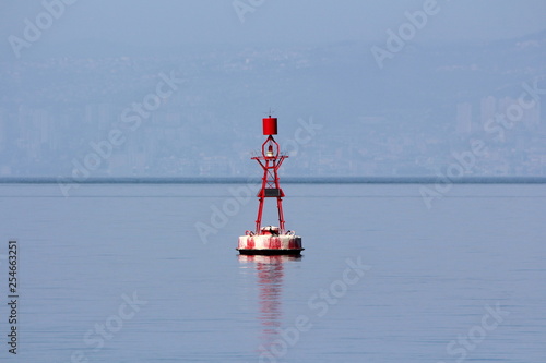 Small red navigational buoy with light on top and solar panels for charging surrounded with calm blue sea on warm spring day