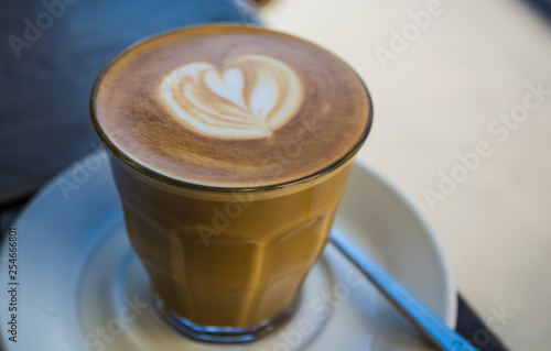 Thick glass tumbler with cappuccino and latte-art in the form heart. Ffresh coffee with milk. On a wooden table with saucer, spoon
