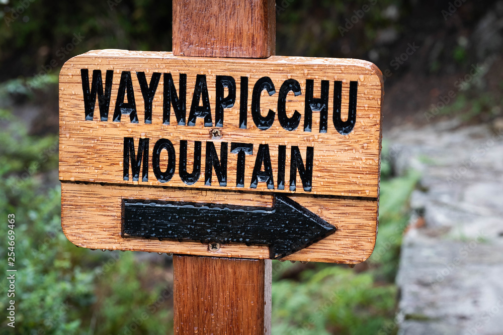 Closeup of wooden pointing sign with black label to Waynapicchu mountain coved with drops