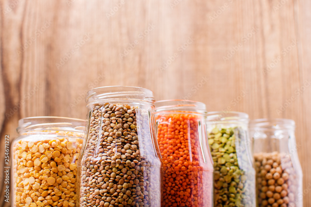 Different legumes in glass jars (chickpeas, green peas, red lentils, Canadian lentils, black lentils, green lentils, yellow peas, red beans) on a wooden background Different legumes in glass jars (chickpeas, green peas, red lentils, Canadian lentils, black lentils, green lentils, yellow peas, red beans) on a wooden background