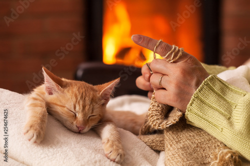 Woman knit in front of the fireplace with her exhausted kitten sleeping in her lap