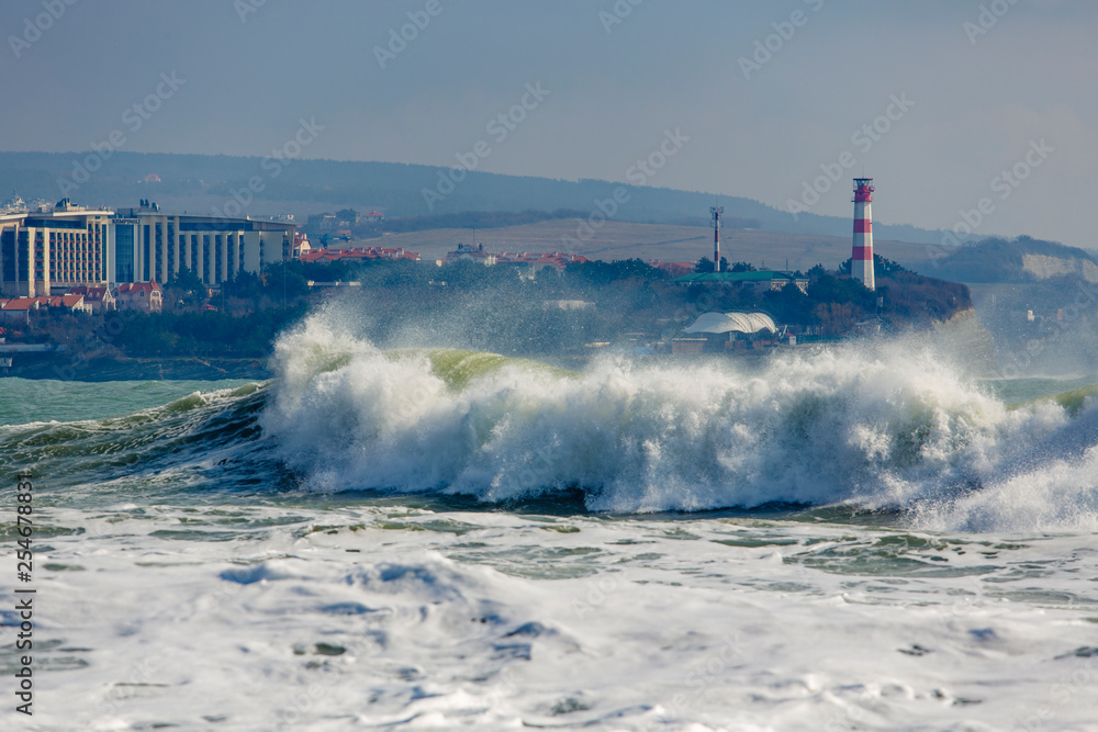 Strong and dangerous storm in the Black sea. Beautiful and big storm wave in Gelendzhik Bay on the background of the resort of Gelendzhik, waterfront and lighthouse. In the background, hotels