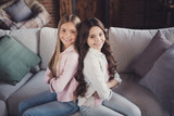 Profile side view portrait of two nice sweet lovely attractive charming cheerful cheery girls sitting on divan back to back folded arms in house loft industrial interior style