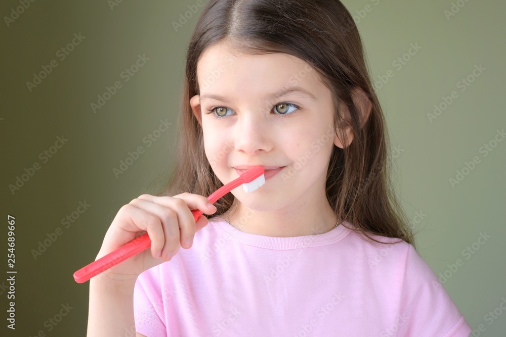 Long haired smiling beautiful caucasian white girl brushing teeth with pink toothbrush on green background.Girl brushing her teeth. Green eyed cute child in violet tshirt doing her morning hygiene. Long haired smiling beautiful caucasian white girl brushing teeth with pink toothbrush on green background.Girl brushing her teeth. Green eyed cute child in violet tshirt doing her morning hygiene.