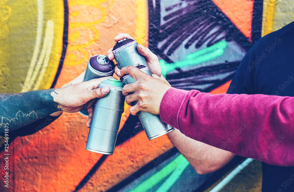 Fototapeta premium Grupa artystów graffiti układających ręce, trzymając puszkę farby w sprayu na tle muralu - Młody malarz przy pracy - Koncepcja sztuki współczesnej, sztuki ulicznej i życia młodzieży