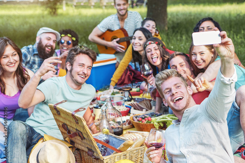 Group of diverse friends taking photo selfie while doing picnic in park - Happy young people having with new mobile phone technology and eating outdoor - Millennial, friendship, food and drink concept