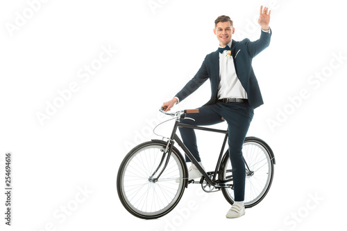 cheerful groom in elegant suit and sneakers sitting on bike and waving hand isolated on white