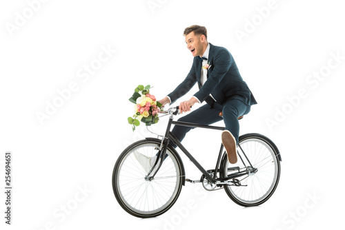 happy groom in elegant suit and sneakers riding bike while holding wedding bouquet isolated on white