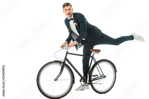 cheerful groom in elegant suit making stunts on bike isolated on white