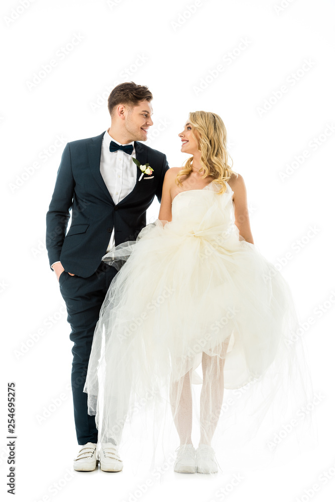 handsome groom and beautiful bride in elegant clothes and sneakers looking at each other isolated on white