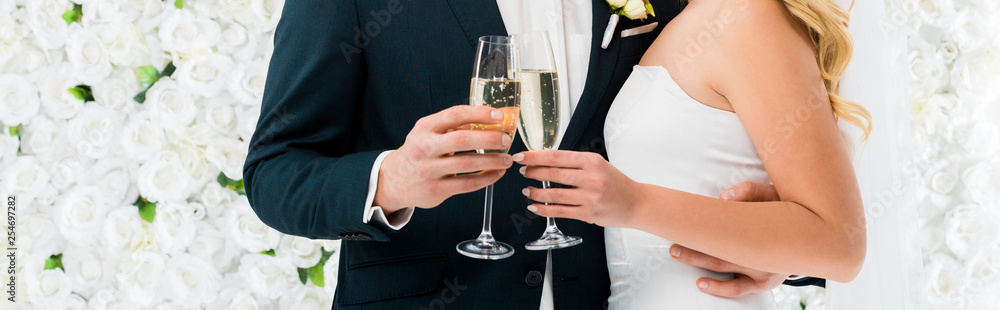panoramic shot of groom and bride holding glasses of champagne on white floral background