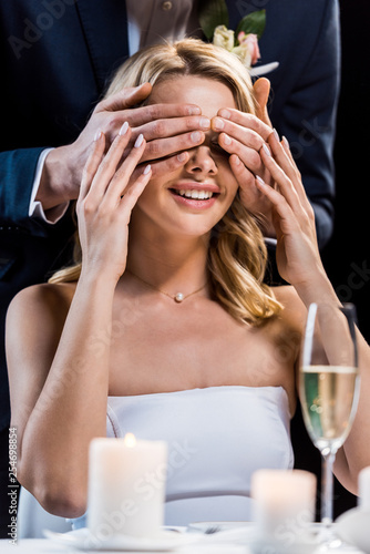 selective focus of happy bride with eyes closed by grooms hands isolated on black