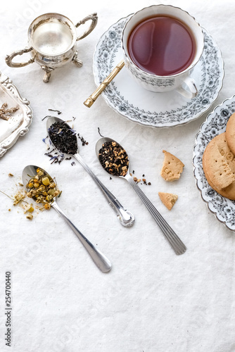 Different types of tea on vintage metal spoons on white fabric background, teacup, dried flower, silver plate, cookies. Vintage food and drink setting styling. Organic healthy well-being lifestyle.