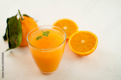 An inviting glass full of orange juice with a fresh mint leaf that floats. In the background an orange divided in two and a whole one with leaves on a white background
