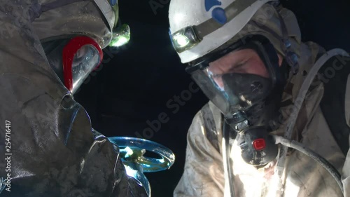 Man in hazmat outfit taking things for laboratory. Two chemists spending time in abandoned factory. Indoors.
