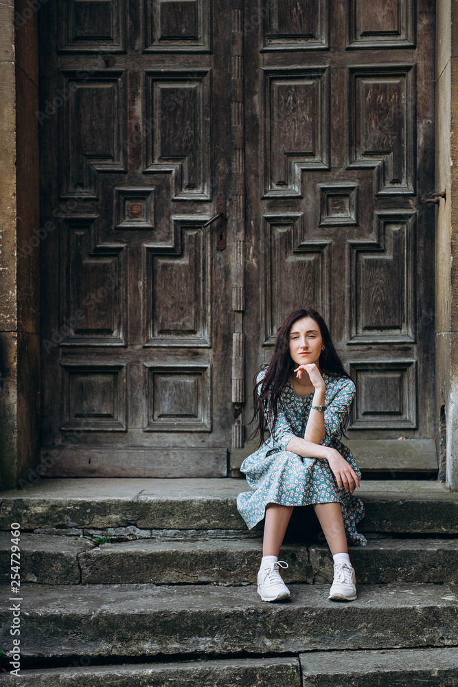 Attractive young woman sitting near old doors in cute summer dress with flower print Attractive young woman sitting near old doors in cute summer dress with flower print