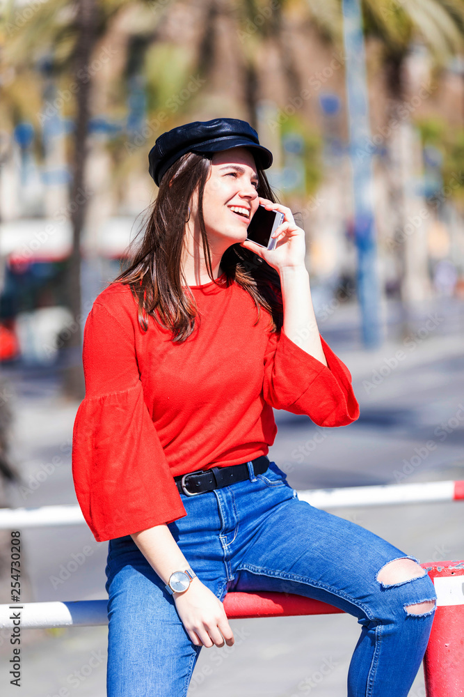 Front view of a beautiful young woman wearing urban clothes sitting on a metallic fence while using a mobile phone outdoors in the street in a bright day
