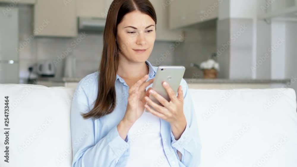 Young brunette woman with brown eyes sitting on sofa in living room using smartphone. Female housewife browsing on mobile phone chatting online with friends. Kitchen on background Young brunette woman with brown eyes sitting on sofa in living room using smartphone. Female housewife browsing on mobile phone chatting online with friends. Kitchen on background