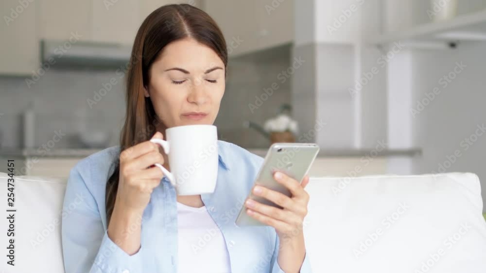 Brunette woman with brown eyes sitting in living room using smartphone. Young female browsing on mobile phone chatting online with friends. Housewife drink coffee from white cup. Kitchen on background Brunette woman with brown eyes sitting in living room using smartphone. Young female browsing on mobile phone chatting online with friends. Housewife drink coffee from white cup. Kitchen on background