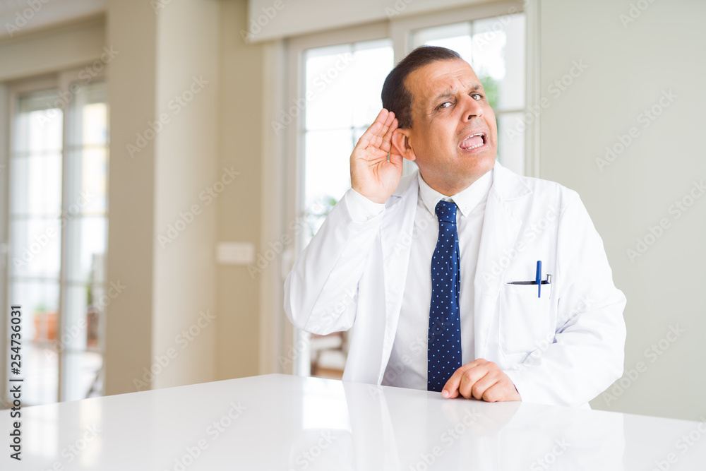 Middle age doctor man wearing medical coat at the clinic smiling with hand over ear listening an hearing to rumor or gossip. Deafness concept.