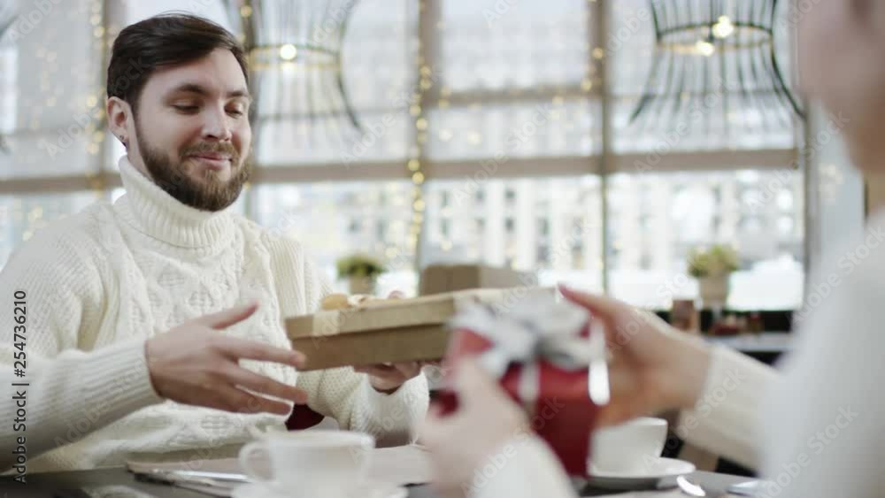 Happy couple exchanging gifts by table and looking inside them and man kissing feomale hand Happy couple exchanging gifts by table and looking inside them and man kissing feomale hand
