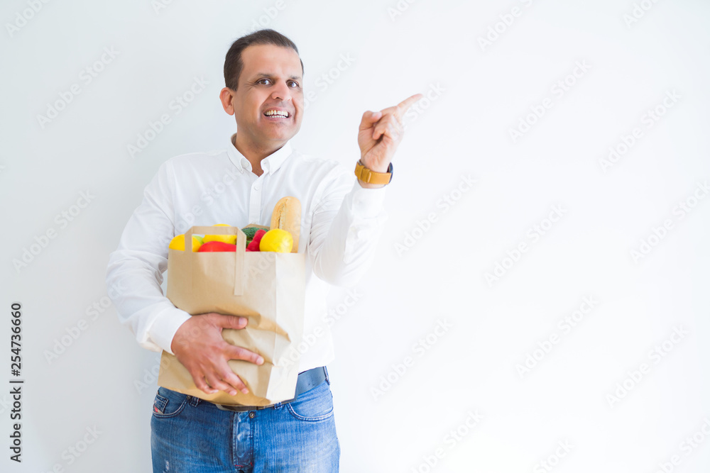 Middle age man holding groceries shopping bag over white background very happy pointing with hand and finger to the side Middle age man holding groceries shopping bag over white background very happy pointing with hand and finger to the side