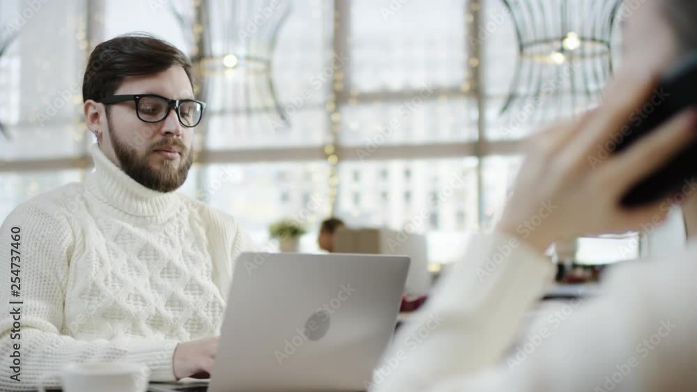 Adult man works on laptop sitting in a cafe with his girlfriend while she is talking over smartphone Adult man works on laptop sitting in a cafe with his girlfriend while she is talking over smartphone