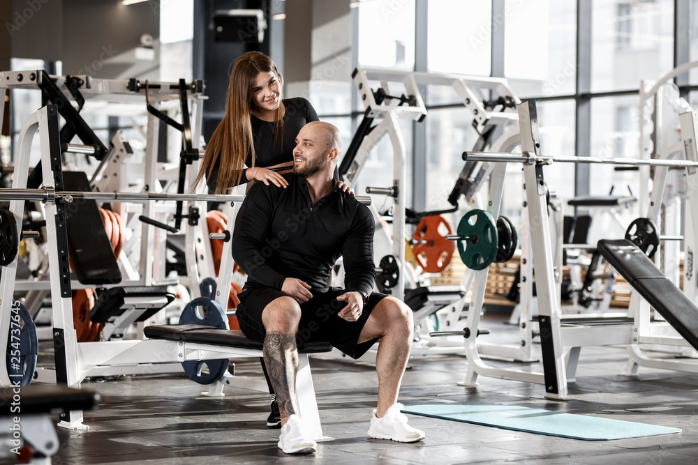 Young slender pretty girl stands behind the athletic man sitting on the spot bench and holds her hands on his shoulders in the modern gym Young slender pretty girl stands behind the athletic man sitting on the spot bench and holds her hands on his shoulders in the modern gym