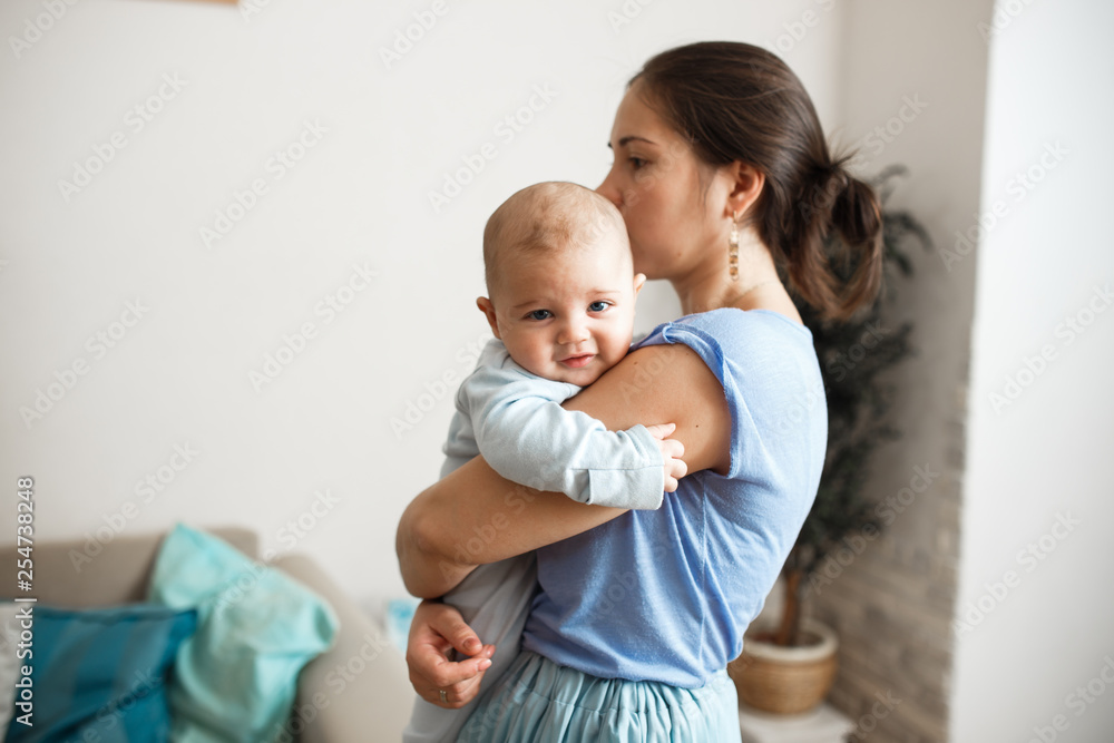 Young mother dressed in light blue t-shirt and skirt is holding her tiny son on her arms in the room at home Young mother dressed in light blue t-shirt and skirt is holding her tiny son on her arms in the room at home