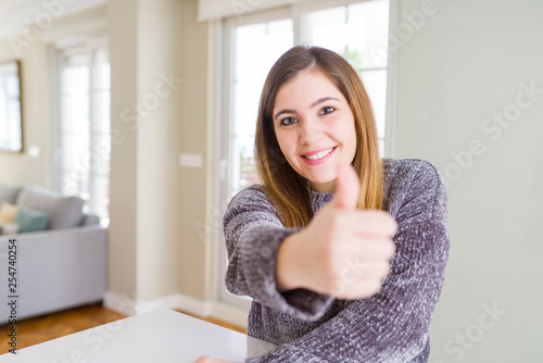 Beautiful young woman at home doing happy thumbs up gesture with hand. Approving expression looking at the camera showing success.