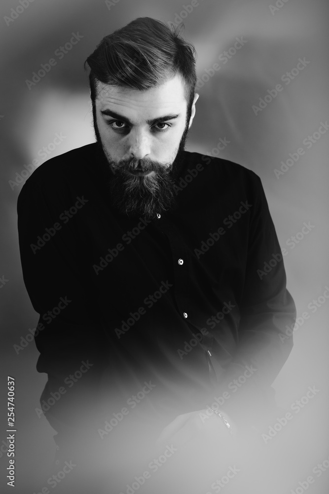 Obraz premium Black and white photo of a stylish man with a beard and stylish hairdo dressed in the black shirt stands in the fog