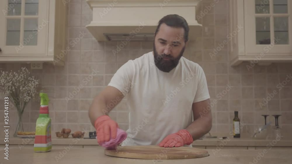 Young man with hipster beautiful beard cleaning the wooden desk and table in gloves with detergent in his new modern house Young man with hipster beautiful beard cleaning the wooden desk and table in gloves with detergent in his new modern house