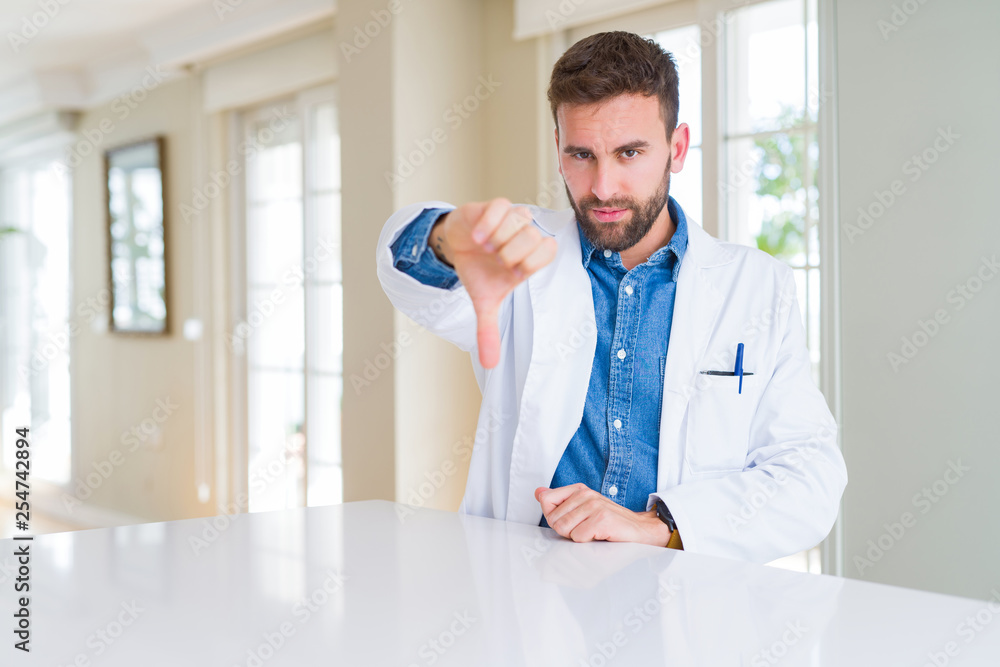 Handsome doctor man wearing medical coat at the clinic looking unhappy and angry showing rejection and negative with thumbs down gesture. Bad expression.